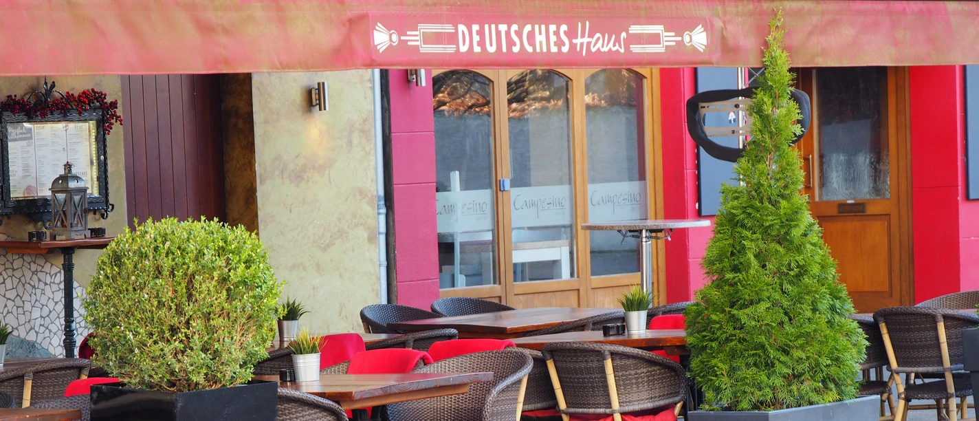 Außenterrasse eines Restaurants mit roten Sitzkissen, Holztischen und Pflanzen in modernen Pflanzkübeln.