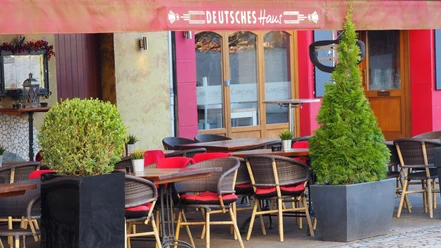 Deutsches Haus Außenterrasse eines Restaurants mit roten Sitzkissen, Holztischen und Pflanzen in modernen Pflanzkübeln.