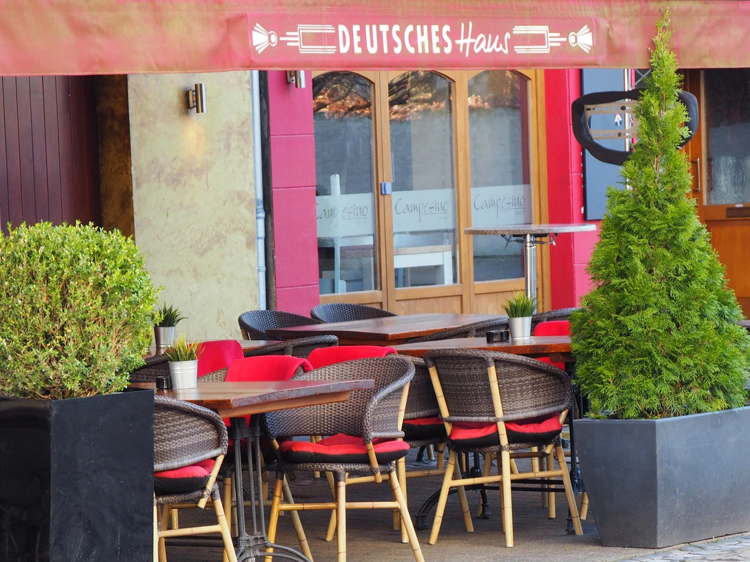Deutsches Haus Außenterrasse eines Restaurants mit roten Sitzkissen, Holztischen und Pflanzen in modernen Pflanzkübeln.