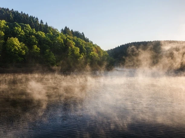Die idyllische Jubachtalsperre Auf dem Wasser der Jubachtalsperre befindet sich Nebel, der leicht braun wirkt. Im Hintergrund Wald und darüber hellblauer Himmel.