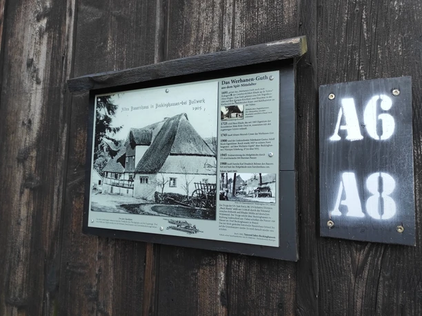 Kleine Infotafel entlang des Weges Eine Info-Tafel an einer Holzwand. Rechts davon eine Tafel mit den Hinweisen auf die Wanderwege A6 und A8.