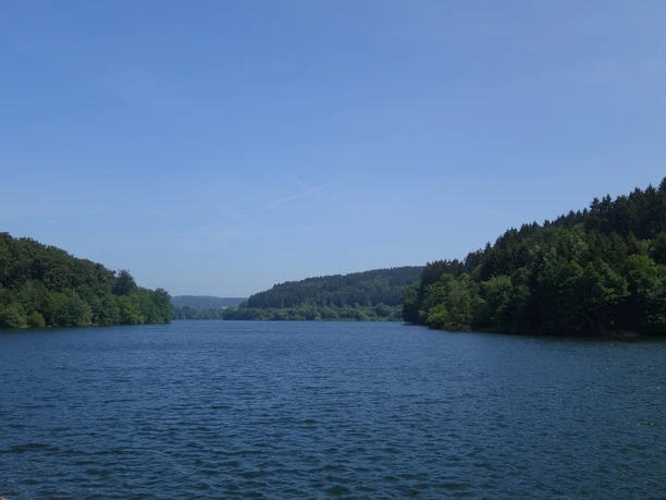 Aussicht von der Staumauer auf die Lingesetalsperre Wasser der Lingesetalsperre umrahmt von Wald. Strahlend blauer Himmel am Horizont.
