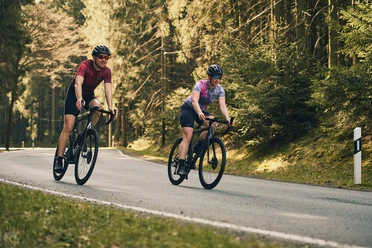 Zwei Rennradfahrer mit Helmen fahren auf einer Straße durch den sonnigen Wald.