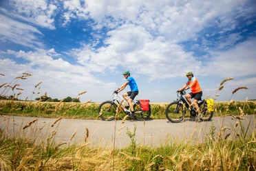 Zwei Radfahrer auf Landstraße zwischen Feldern unter weitem Wolkenhimmel