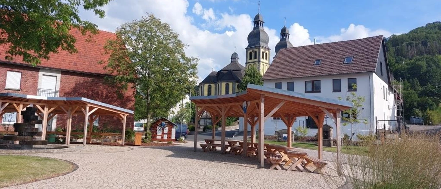 Dorfplatz in Padberg mit Holzdächern, Bänken und Kirche vor bewaldetem Hang