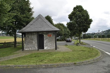 Kleine Kapelle mit Schieferdach an einer Straße, umgeben von Bäumen und Feldern.