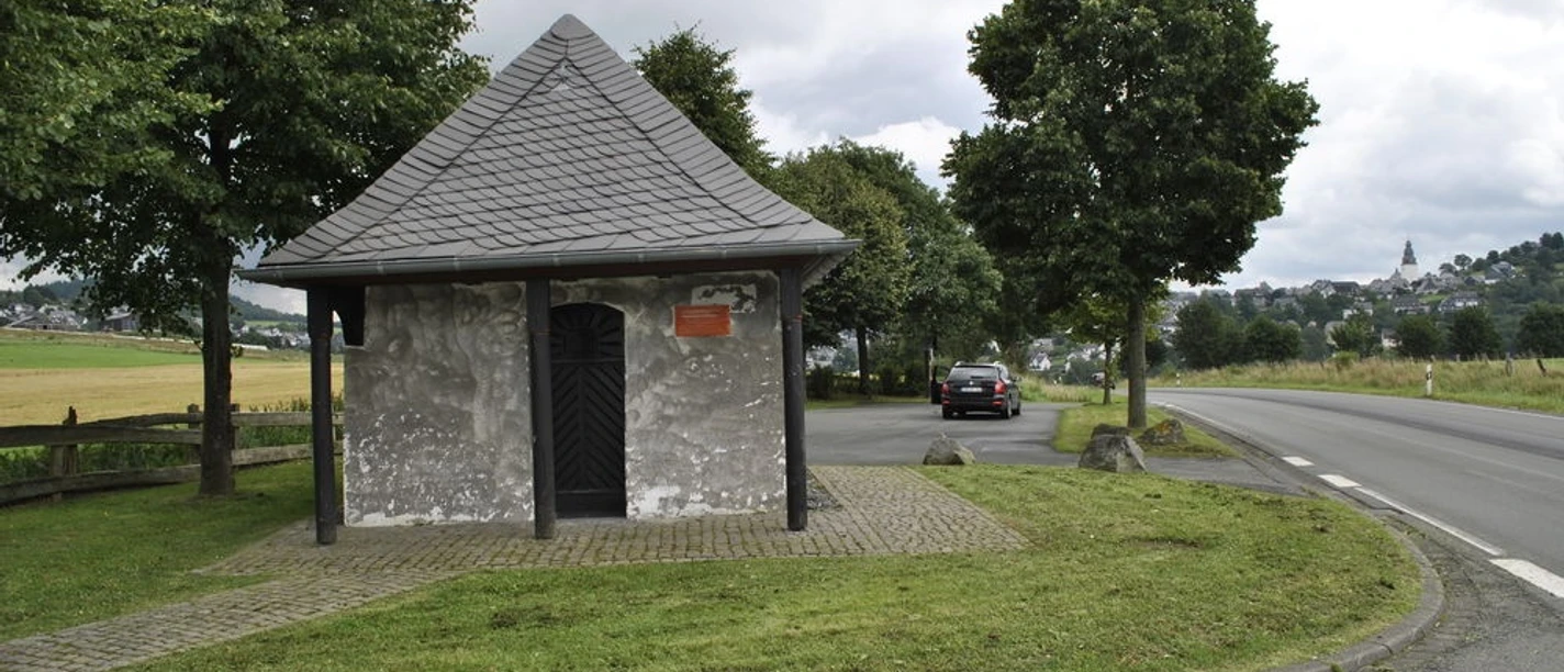 Kleine Kapelle mit Schieferdach an einer Straße, umgeben von Bäumen und Feldern.