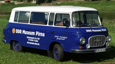 Blauer Oldtimer-Bus mit weißem Dach, beschriftet mit "DDR Museum Pirna", steht auf einer grünen Wiese.