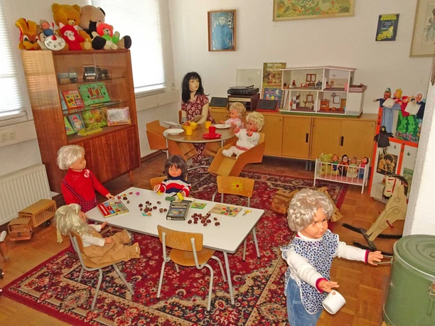 Kinderkrippe DDR-Museum Pirna Ein Raum voller Puppen in verschiedenen Posen; einige sitzen an einem Tisch mit Spielzeug, andere an einem runden Tisch, umgeben von Regalen mit Büchern und Stofftieren.