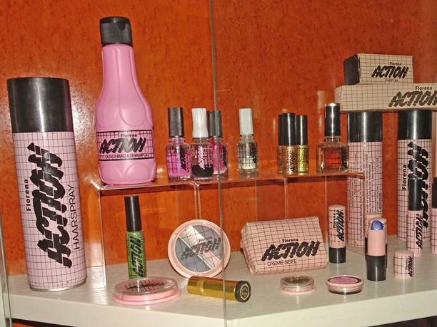 Kosmetikartikel im DDR-Museum Pirna Kosmetikprodukte der Marke Florena Action in einem Regal, darunter Haarspray, Duschgel, Nagellacke und Seife, vor einem orangefarbenen Hintergrund.