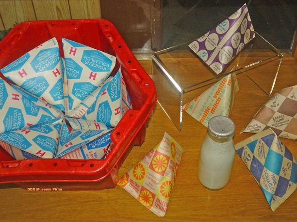 Rode krat met blauwe en witte gecondenseerde melkpakken, daarnaast verschillende gekleurde melkpakken en een glazen fles op een houten tafel.