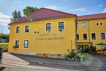 Scheidehaus am "Marie Louise Stolln" Gelbes Gebäude in traditionellem Stil mit rotem Ziegeldach, umgeben von blühenden Blumen und Gehweg.
