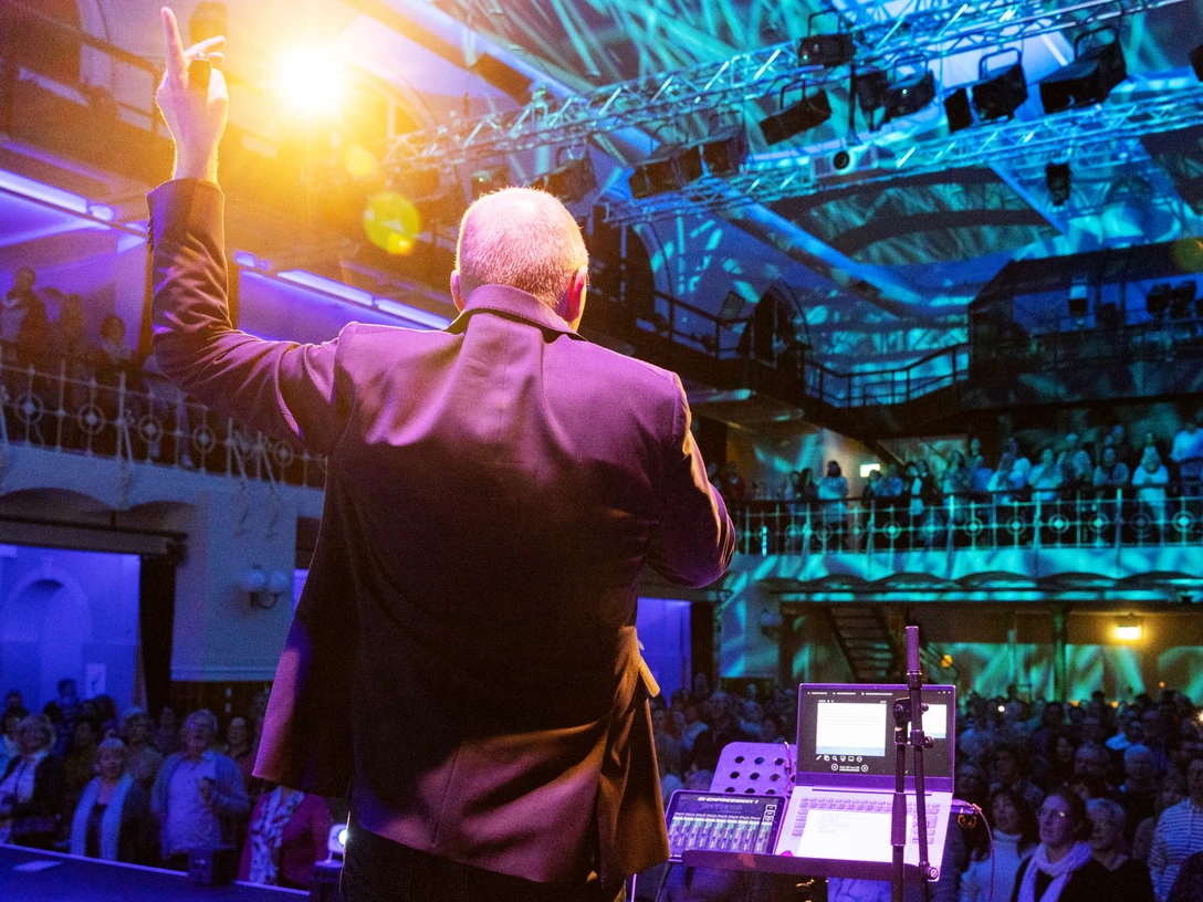 RUDELSINGEN_2023_Live-c-Rudelsingen GmbH_Joachim_Pantel.jpg Ein Bühnensprecher steht vor einem Publikum in einer beleuchteten Veranstaltungshalle.