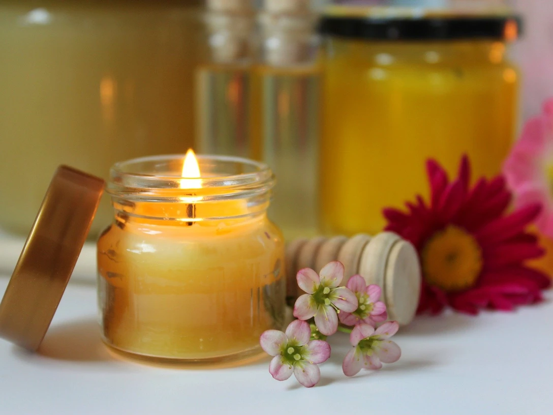 Bienenwachs-Kerzenwerkstatt-2.jpg Glas mit brennender Bienenwachskerze, umgeben von Honiggläsern und bunten Blumenarrangements.