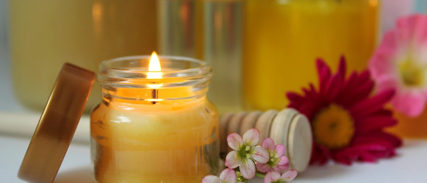 Bienenwachs-Kerzenwerkstatt-2.jpg Glas mit brennender Bienenwachskerze, umgeben von Honiggläsern und bunten Blumenarrangements.