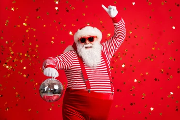 Photo of retired old man grey beard hold vintage disco ball dance funky wear santa x-mas costume suspenders sunglass gloves striped shirt cap isolated red color background Santa Claus holds a disco ball in his hand and stands in front of a red wall