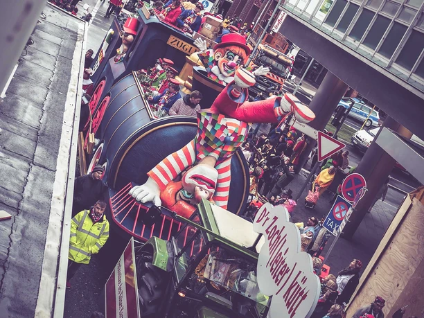 Karneval_1.jpg Ein bunter Festwagen mit einem riesigen Clown und Radfahrern während eines Karnevalsumzugs.