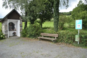 Kleiner Schotterparkplatz mit Bank, Wegkapelle und Parkschild in grüner Umgebung.