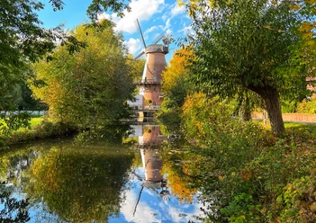 Eine historische Mühle mit drehenden Flügeln spiegelt sich in einem von buntem Herbstlaub umsäumten Fluss.