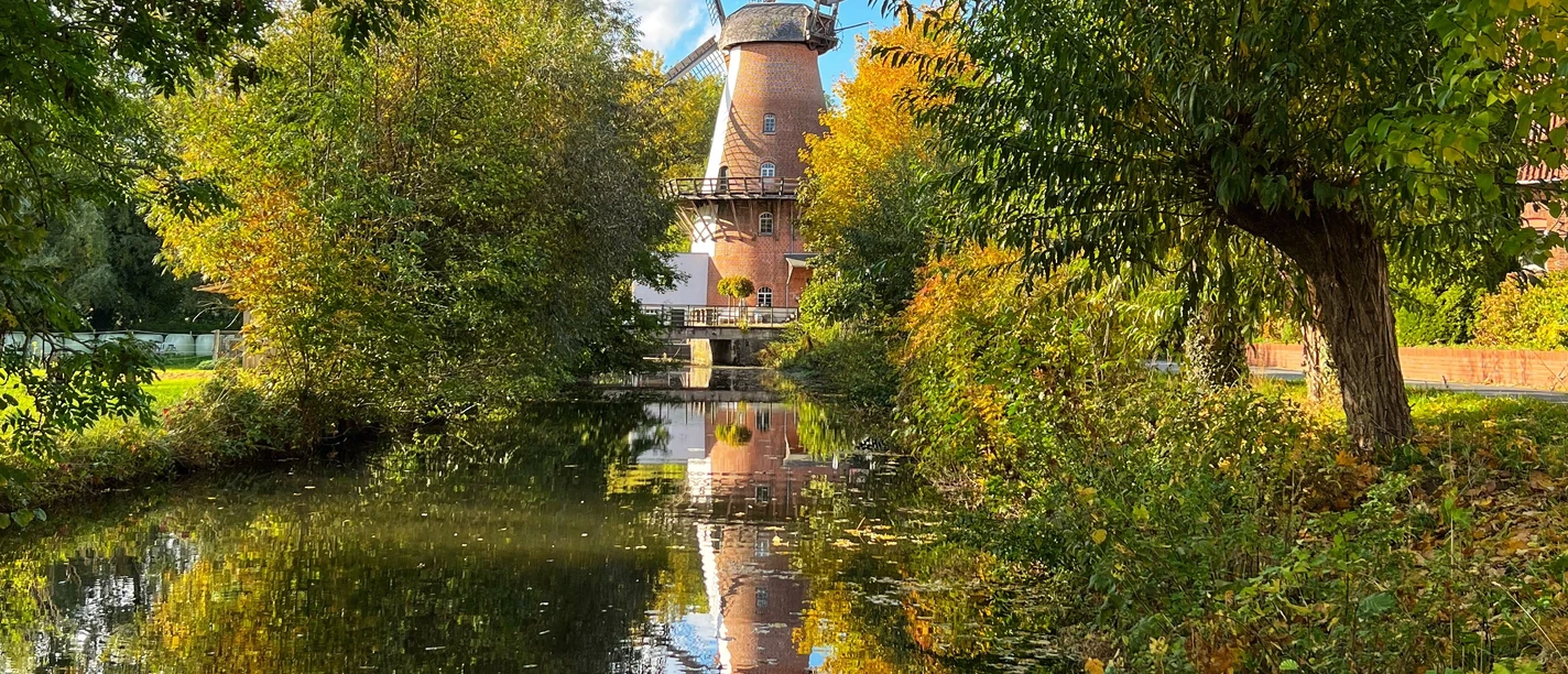 Eine historische Mühle mit drehenden Flügeln spiegelt sich in einem von buntem Herbstlaub umsäumten Fluss.