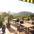 Panoramablick von der Hotel-Restaurant Terrasse auf grüne Hügel mit leeren Tischen im Sonnenlicht.