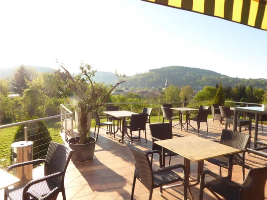 Panoramablick von der Hotel-Restaurant Terrasse auf grüne Hügel mit leeren Tischen im Sonnenlicht.