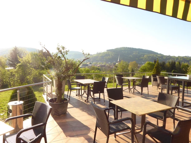 Panoramablick von der Hotel-Restaurant Terrasse auf grüne Hügel mit leeren Tischen im Sonnenlicht.