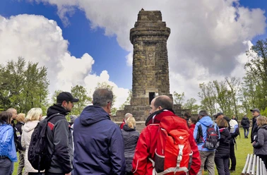 Zahlreiche Menschen stehen vor einem historischen Steinturm, umgeben von Bäumen unter wolkigem Himmel.