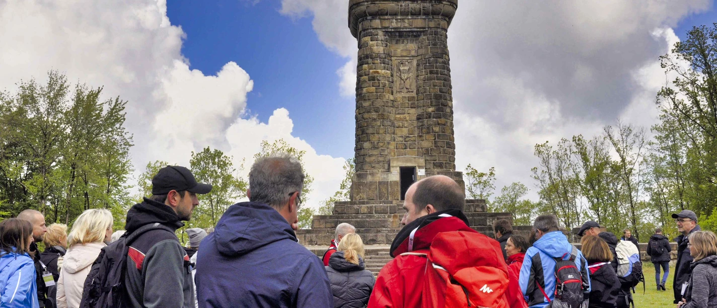 Zahlreiche Menschen stehen vor einem historischen Steinturm, umgeben von Bäumen unter wolkigem Himmel.