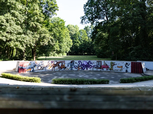Amphitheater im Grünen mit bunten Graffitis auf der Rückwand, umgeben von Bäumen bei Sonnenschein.