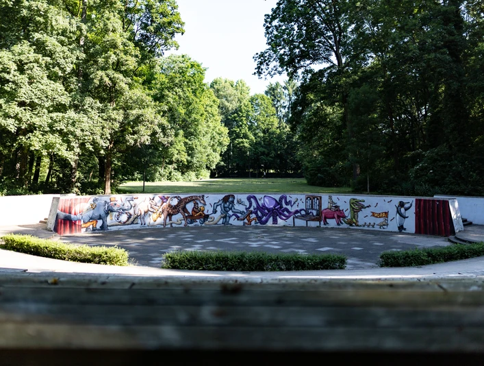 Amphitheater im Grünen mit bunten Graffitis auf der Rückwand, umgeben von Bäumen bei Sonnenschein.