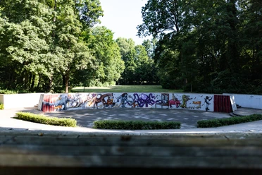 Mohns Park Amphitheater im Grünen mit bunten Graffitis auf der Rückwand, umgeben von Bäumen bei Sonnenschein.
