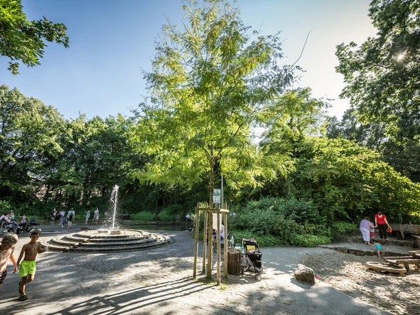 Ein grüner Spielplatz mit Springbrunnen, umgeben von Bäumen, spielenden Kindern und Erwachsenen.