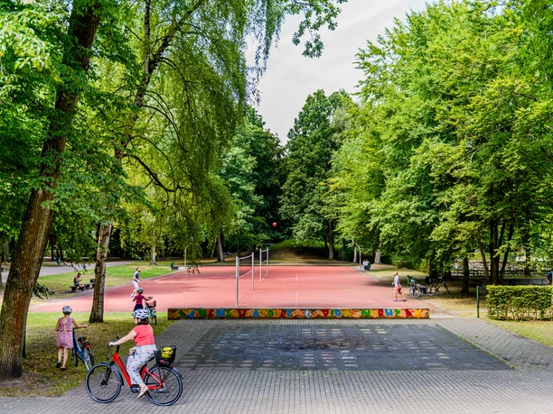 Mohnspark. Gütersloh Ein weitläufiger Park mit einem roten Volleyballfeld, umgeben von üppigem Grün und Spaziergängern.
