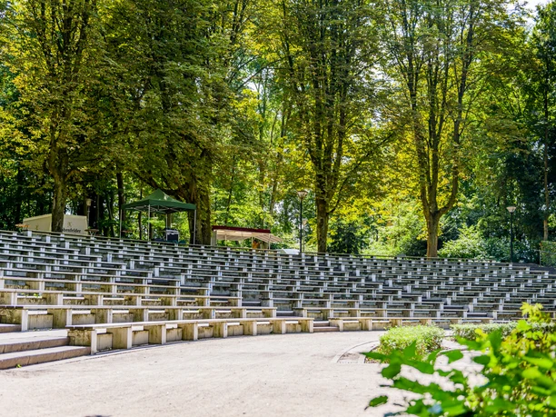 Ein amphitheaterartiger Sitzbereich mit leeren Stufenbänken unter hohen, schattenspendenden Bäumen.