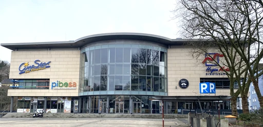 CineStar Hagen©Tim Lichtenberg.jpeg Ein modernes Gebäude mit breiter Glasfront und mehreren Geschäftsschildern an der Fassade.