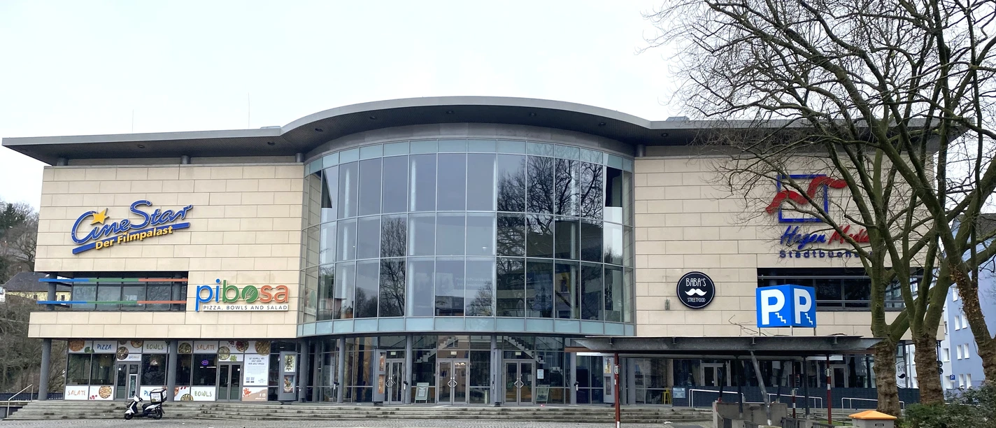 CineStar Hagen©Tim Lichtenberg.jpeg Ein modernes Gebäude mit breiter Glasfront und mehreren Geschäftsschildern an der Fassade.