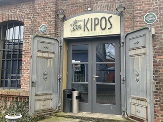 Eingang zum griechischen Restaurant Haus Kipos mit geöffneten Holztüren in einem Backsteingebäude.