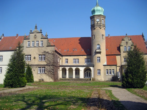 Historisches Schloss mit rotem Ziegeldach, Eckturm mit grünem Dach, umgeben von gepflegtem Rasen.