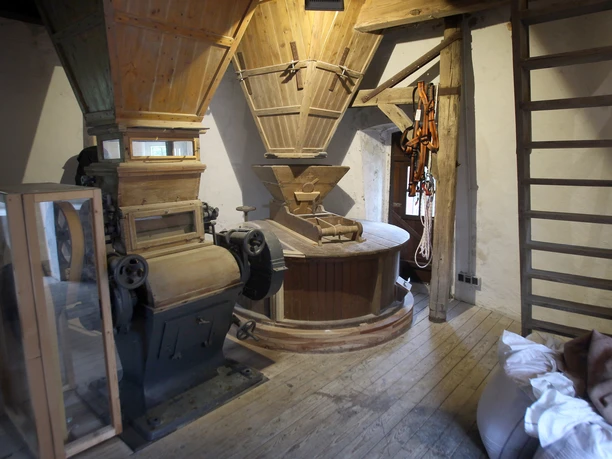 Das Innere einer historischen Windmühle mit hölzernem Mahlwerk und alten Maschinenteilen aus Metall.