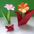 Origami Blumen Origami-Kunst: Zwei Blumen in Grün- und Orangetönen und zwei rote Papierkraniche vor grünem Hintergrund.