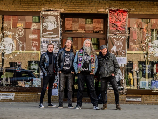 Vier Männer stehen lächelnd vor einem Schallplattenladen, bekleidet mit Lederjacken und Band-Shirts.