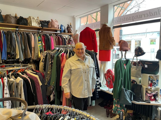 Dombrowsky1.jpg Eine lächelnde Frau steht in einem Second-Hand-Laden umgeben von Kleidung und Accessoires.