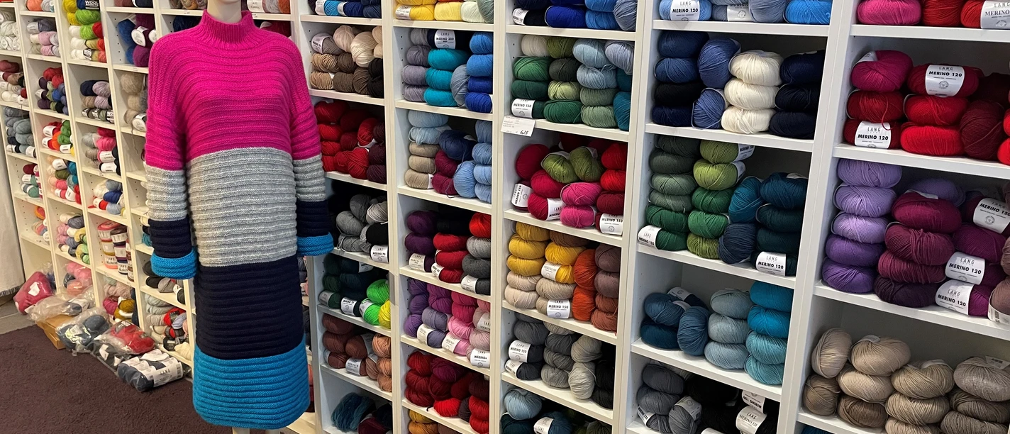 maris.masche1.jpg Eine Schaufensterpuppe im langen, gestreiften Kleid steht vor einem Regal voller bunter Wollknäuel.