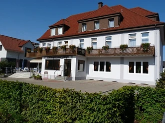 Ein dreistöckiges Gebäude mit rotem Dach, Blumenkästen an den Balkonen, umgeben von einem Garten.
