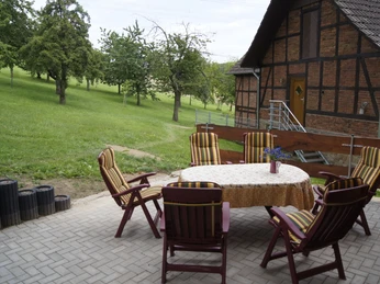 Foto_Ferienwohnung Milan.JPG Sitzgruppe mit Holzmöbeln steht auf gepflasterter Fläche vor rustikalem Fachwerkhaus, umgeben von Bäumen.
