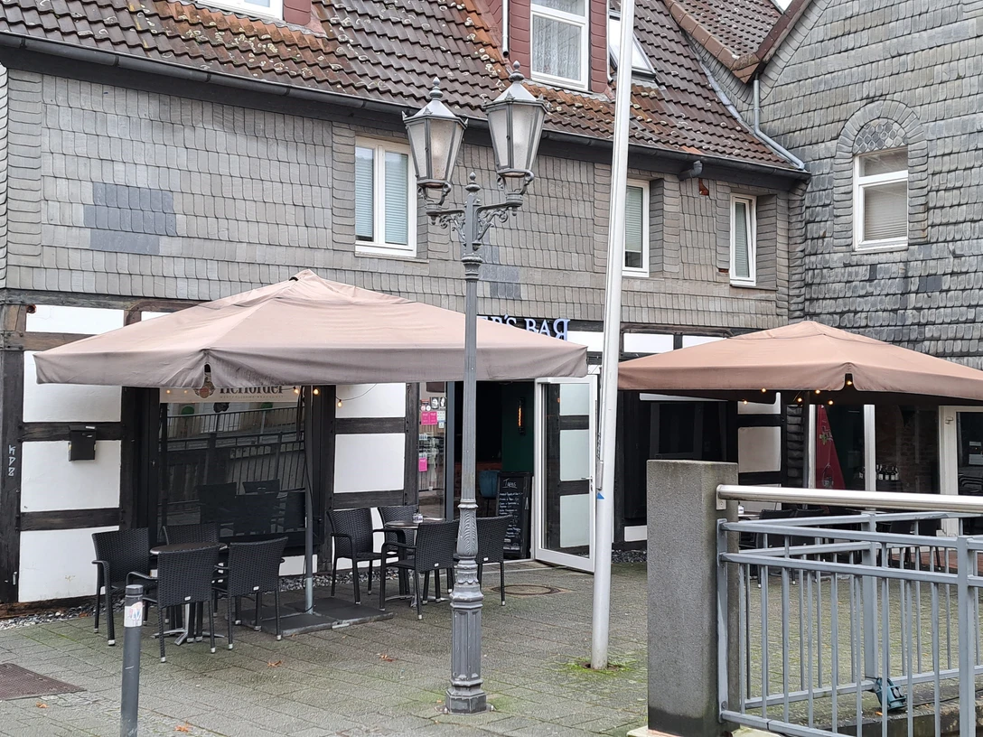 Blick auf ein Fachwerkhaus mit Schindeldach, beidseitig Außenterrassen mit Sonnen- und Regenschirmen.