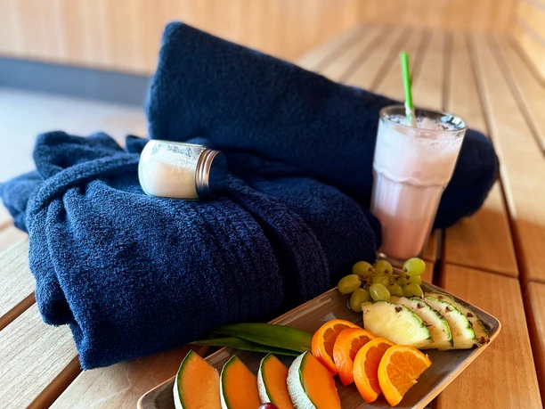 Verwöhn-Paket_Seesauna_b.jpg Wellness-Ambiente mit frischem Obst, Getränk, schwarzen Handtüchern und Creme auf Holztisch.