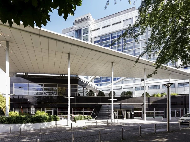 Pullman Cologne 5366 Das Bild zeigt die moderne Frontansicht des Pullman Cologne Hotels mit seiner markanten Architektur und großen Fenstern. Vor dem gepflegten Gebäude erstreckt sich eine überdachte Vorfahrt mit schlanken Säulen, die einen einladenden Eingangsbereich schaffen.