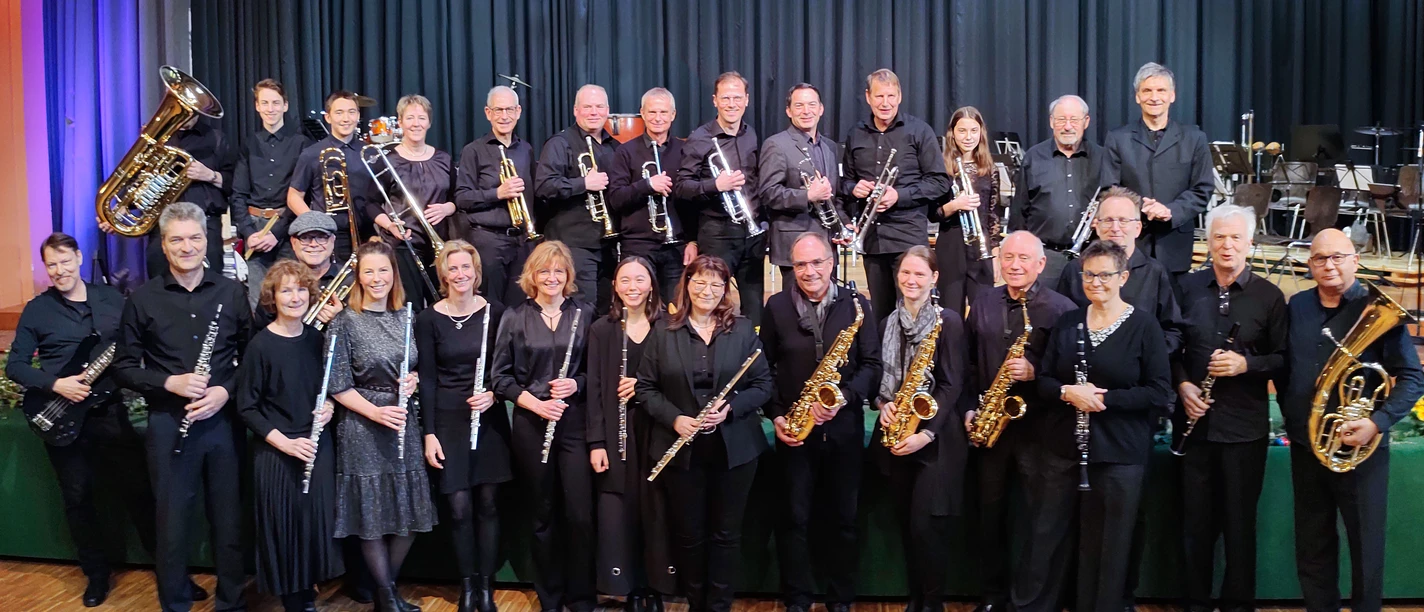 Sülztaler Blasorchester Eine große Musikgruppe in schwarzer Kleidung posiert lächelnd mit ihren Blasinstrumenten auf einer Bühne.